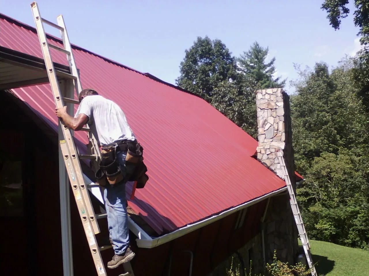 Roofer on a ladder beside a red metal roof and stone chimney — Soffit & Fascia Repair by Roofing Brothers in Larimore